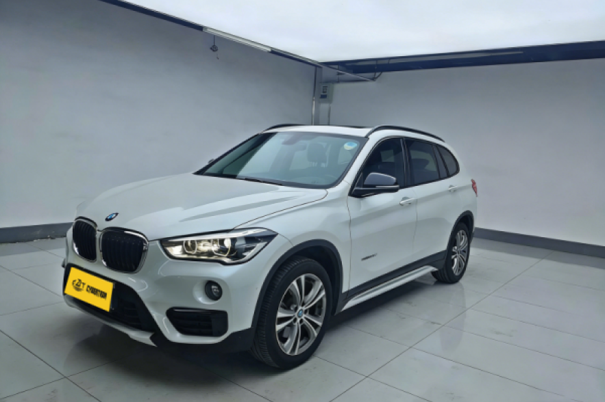 White BMW X1 M Sport SUV in indoor showroom, side-front view with alloy wheels, roof rails and yellow license plate.