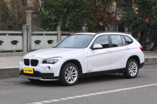White BMW X1 E84 SUV parked on street, side-front view with alloy wheels, yellow plate, trees and stone wall in background.