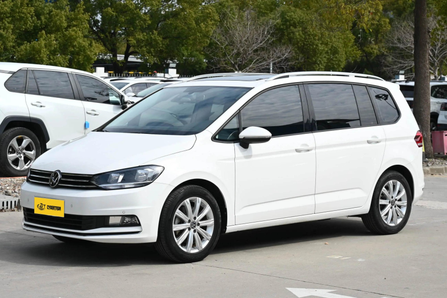 White Volkswagen Touran MPV three-quarter front side view parked on paved parking lot with leafy trees background